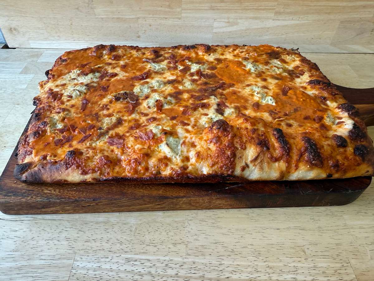 Rectangular pan-style pizza with a thick, crispy crust, generously topped with melted cheese and a creamy orange-hued sauce, served on a wooden paddle.