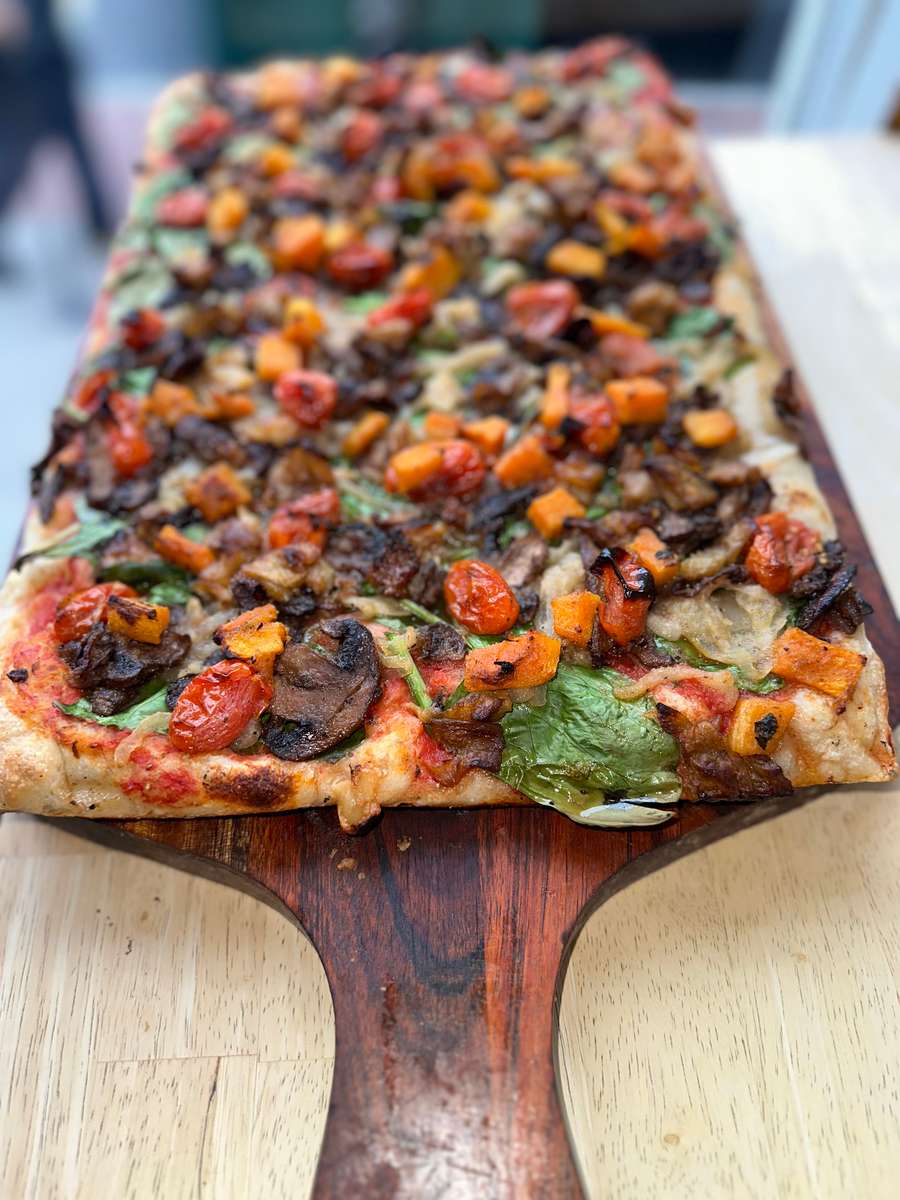 Close-up of a rectangular wood-fired vegetable pizza topped with roasted cherry tomatoes, sautéed mushrooms, caramelized onions, spinach, and golden butternut squash cubes, served on a rustic wooden pizza peel.