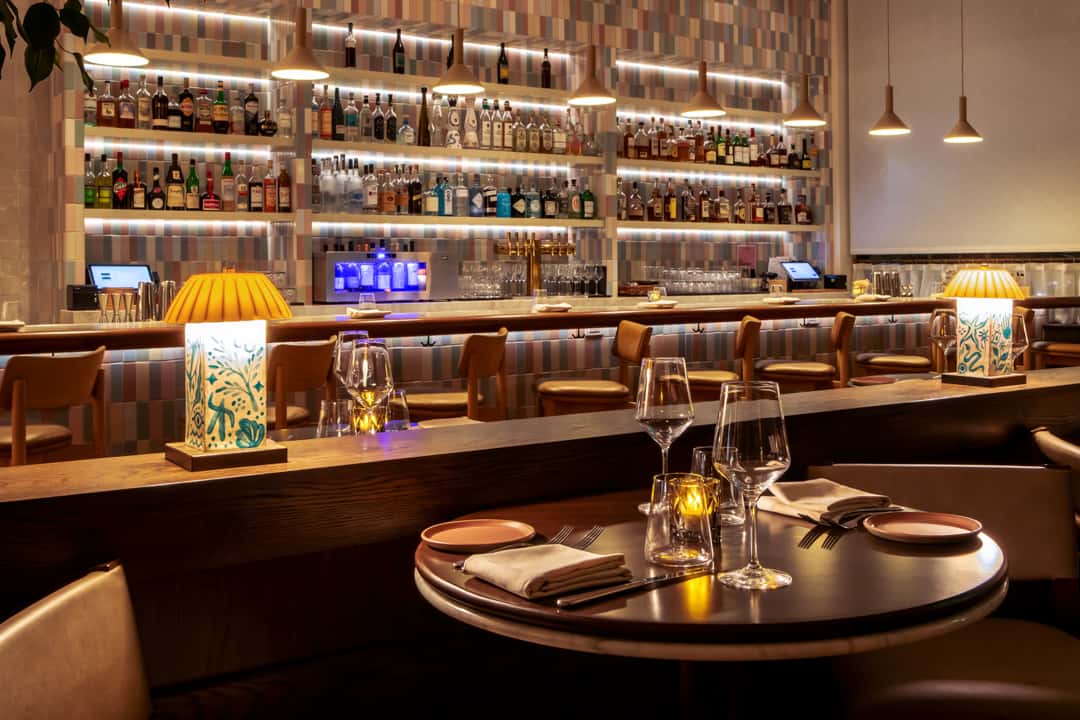 Elegant restaurant bar with illuminated liquor shelves, warm ambient lighting, and intimate dining tables set for upscale cocktails and dinner.