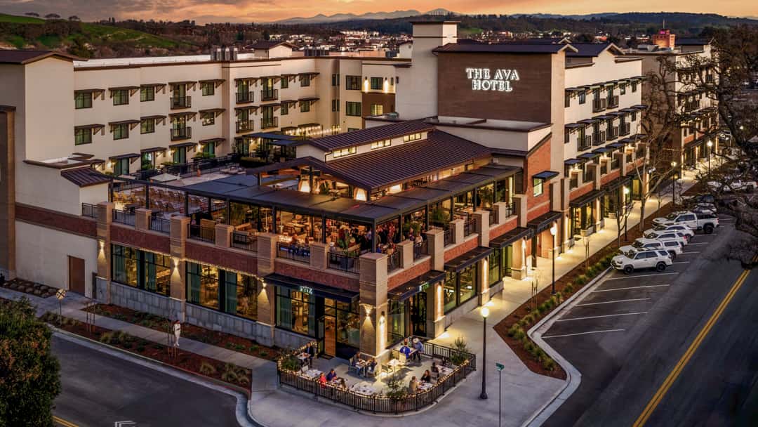 Aerial view of The Ava Hotel featuring a rooftop restaurant, outdoor dining patio, and modern mixed-use development at sunset.