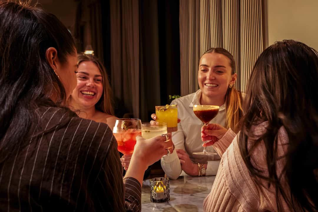 Friends enjoying colorful craft cocktails and conversation in a warm, upscale restaurant setting.