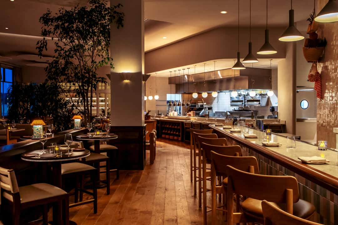 Upscale restaurant interior with open kitchen, marble bar seating, warm pendant lighting, and elegant dining tables.
