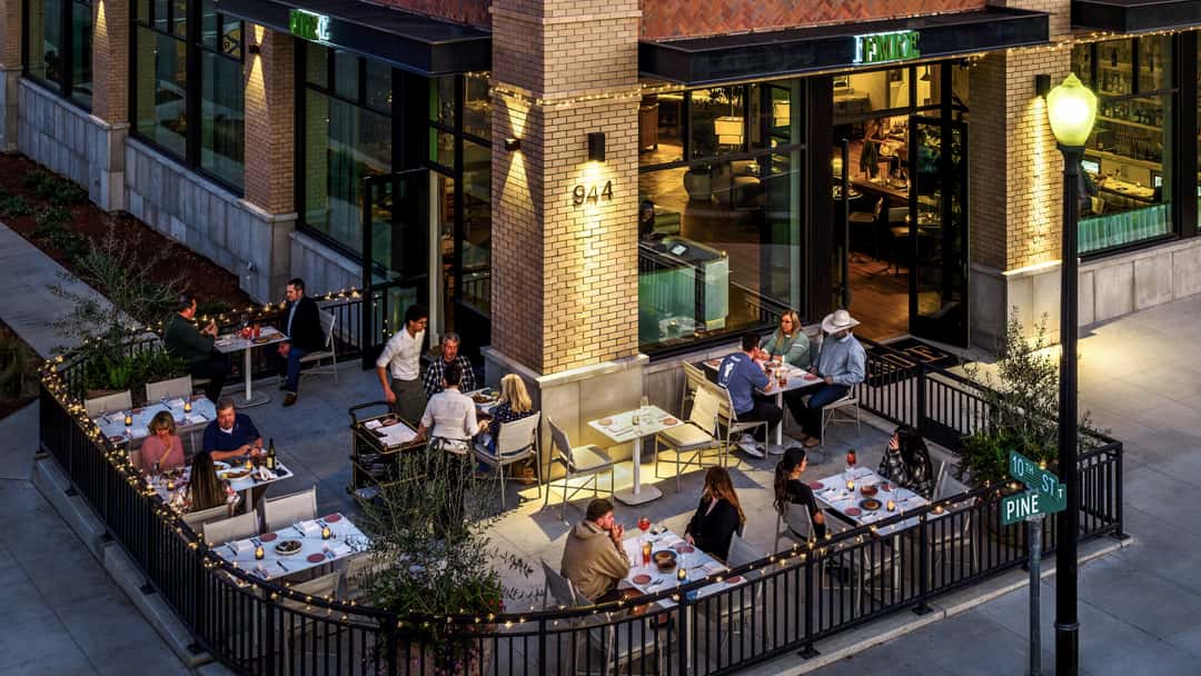 Outdoor restaurant patio with guests dining at candlelit tables beside a modern corner restaurant in the evening.