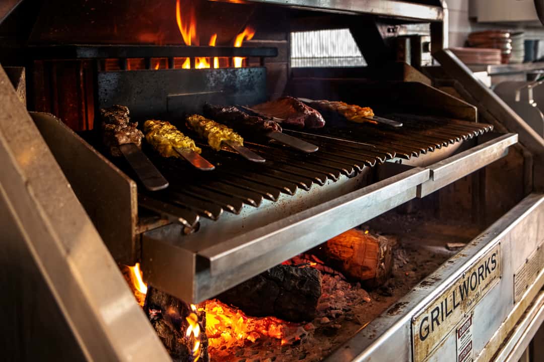 Wood-fired grill cooking skewered meats over open flames, showcasing live-fire grilling and premium restaurant cuisine.