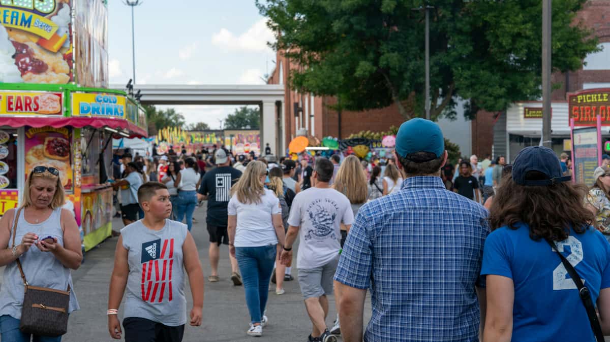 Crowd of people walking through a busy outdoor fair with food stands and colorful signs