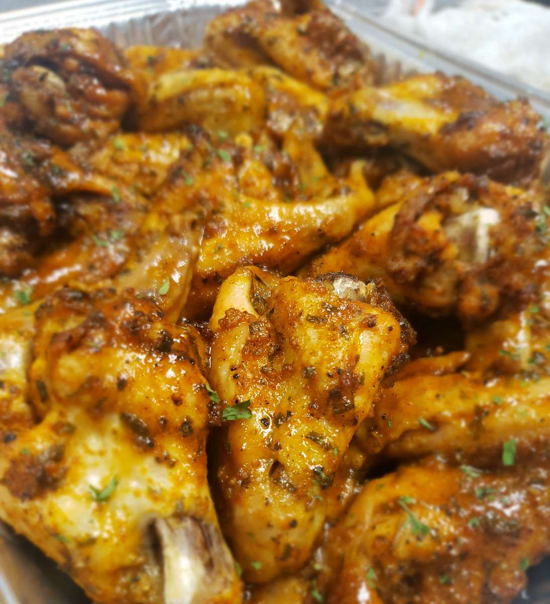 Glossy, golden chicken wings coated in a spicy glaze and herbs, piled high in a catering foil tray.