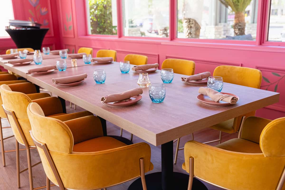Chic restaurant dining setup featuring a long wooden table with mustard yellow velvet chairs, modern pink wall accents, and stylish place settings with beige napkins and blue glassware—perfect for trendy brunch spots, private dining, or upscale event venues.