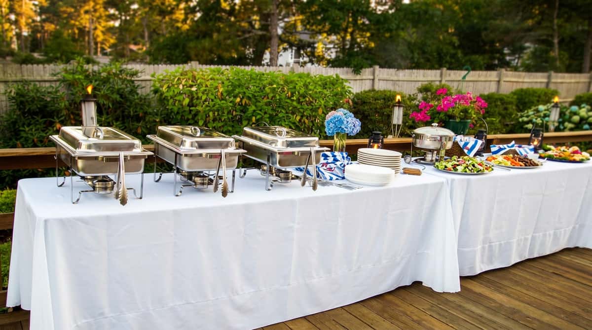 The image shows an elegant outdoor catering setup with a long table covered in a white tablecloth. Chafing dishes are arranged neatly for serving food, surrounded by simple yet tasteful decorations, including floral accents. The setting feels refined and welcoming, perfect for a garden party, wedding, or special event.