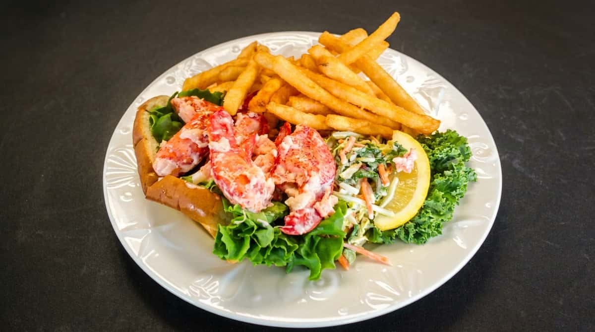 The image features a classic lobster roll served on a bed of fresh greens, paired with a generous portion of crispy golden fries and a lemon wedge. The lobster meat appears tender and lightly dressed, offering a fresh, coastal feel that's both hearty and appetizing.