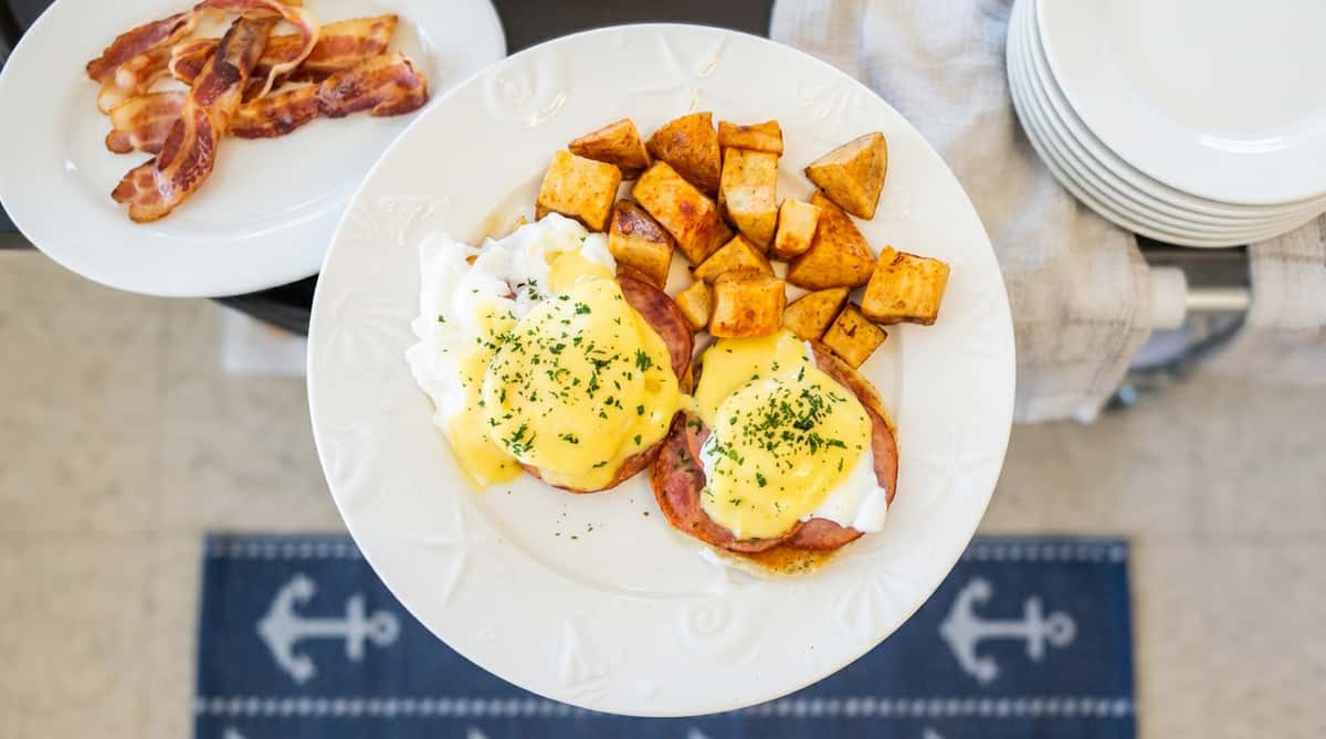 The image shows a well-presented brunch dish featuring two eggs Benedict served on toasted English muffins, topped with creamy hollandaise sauce and a sprinkle of herbs. Alongside, there's a generous portion of crispy, golden-brown breakfast potatoes. The plate is clean and bright, giving the dish a fresh and appetizing, coastal café feel.