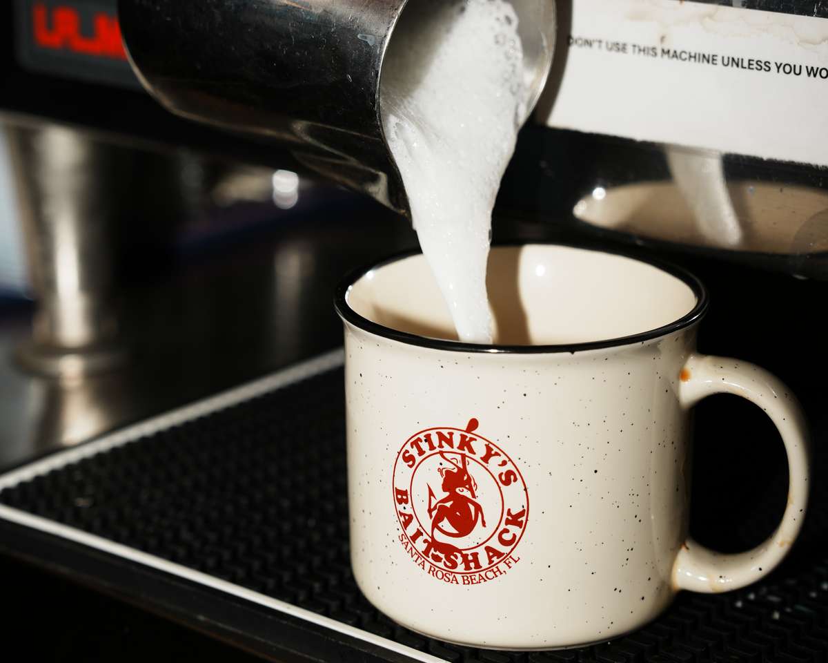 Milk being poured into a Stinky's Bait Shack branded mug at an espresso machine, preparing a hot coffee drink.