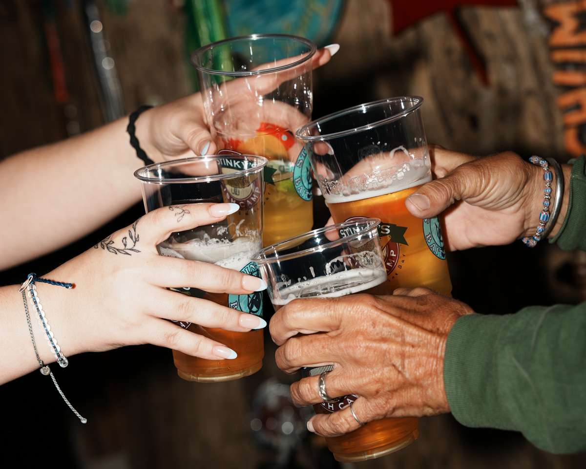 Friends clinking drinks—beer and cocktails—in branded cups at Stinky's Bait Shack, with rustic fishing-themed décor in the background.