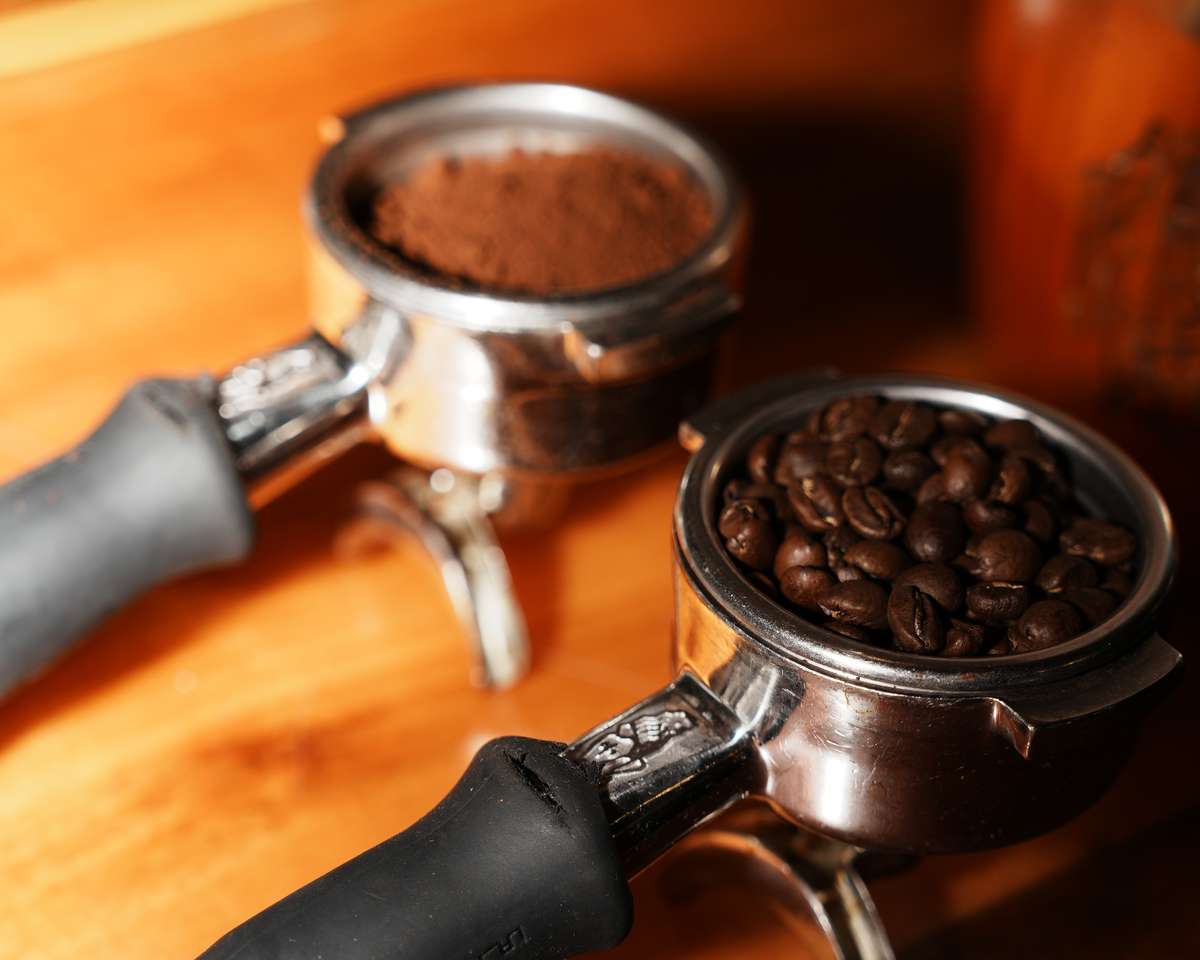 Two espresso portafilters—one filled with freshly ground coffee and the other with whole coffee beans—resting on a wooden counter, ready for brewing.