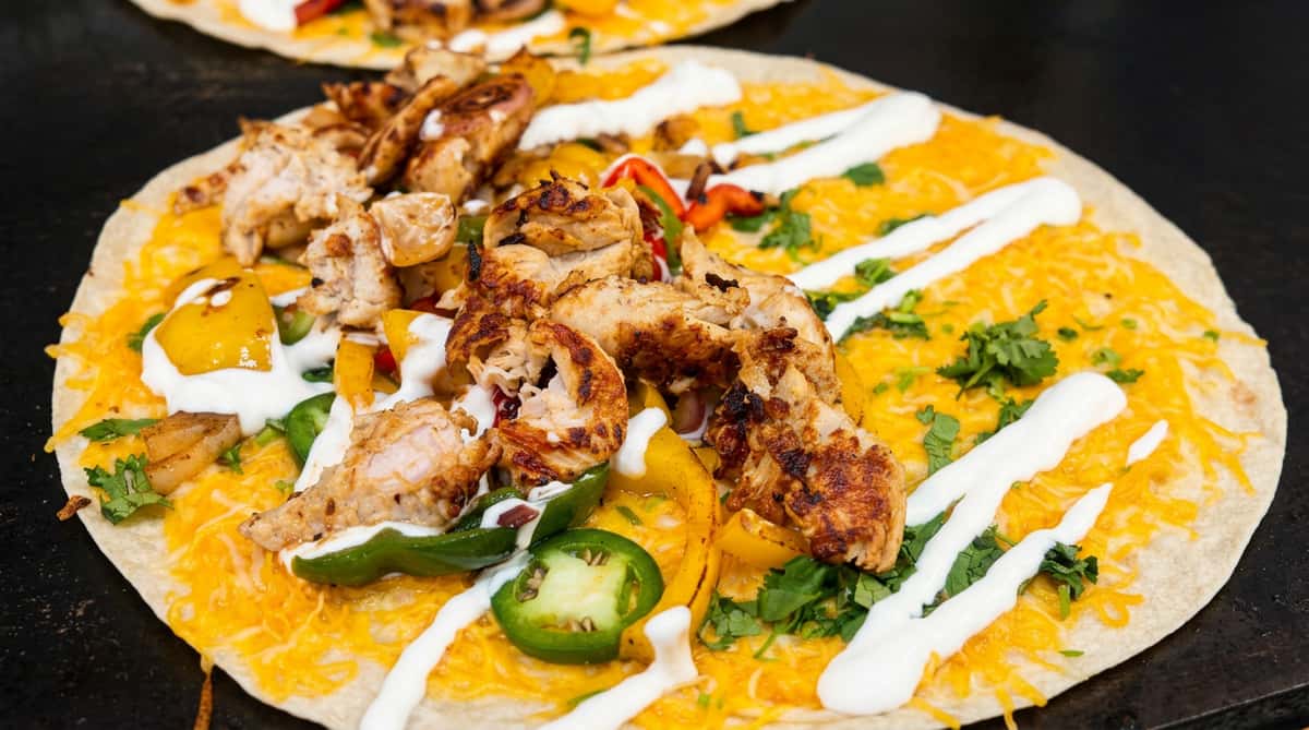 Chicken fajita quesadilla with melted orange cheese, grilled chicken strips, bell peppers, onions, jalapenos, cilantro, and white sour cream drizzle on a large tortilla.