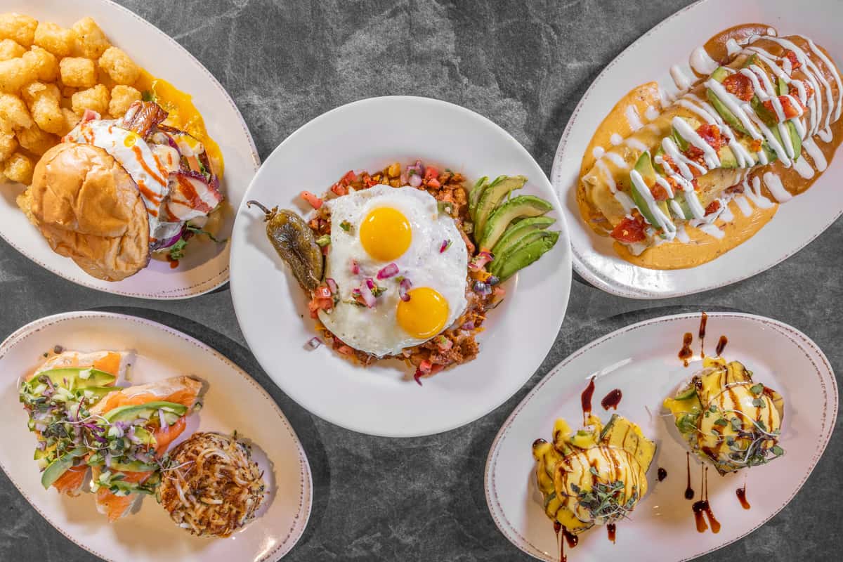 Overhead view of assorted breakfast dishes on white plates, featuring sunny-side-up eggs, fresh greens, roasted vegetables, and colorful brunch favorites arranged on a dark table.