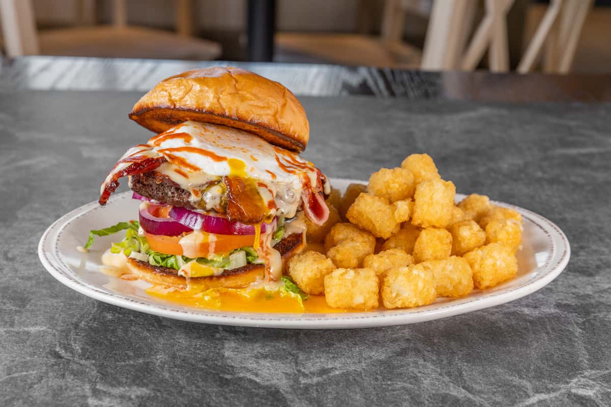 Juicy cheeseburger stacked with lettuce, tomato, red onion, melted cheese, and a toasted sesame seed bun, served with crispy golden tater tots on a white plate.