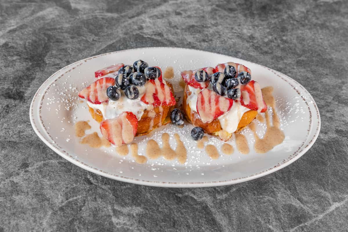 Two servings of golden French toast topped with fresh strawberries, blueberries, and a generous drizzle of white cream, served on a white plate.