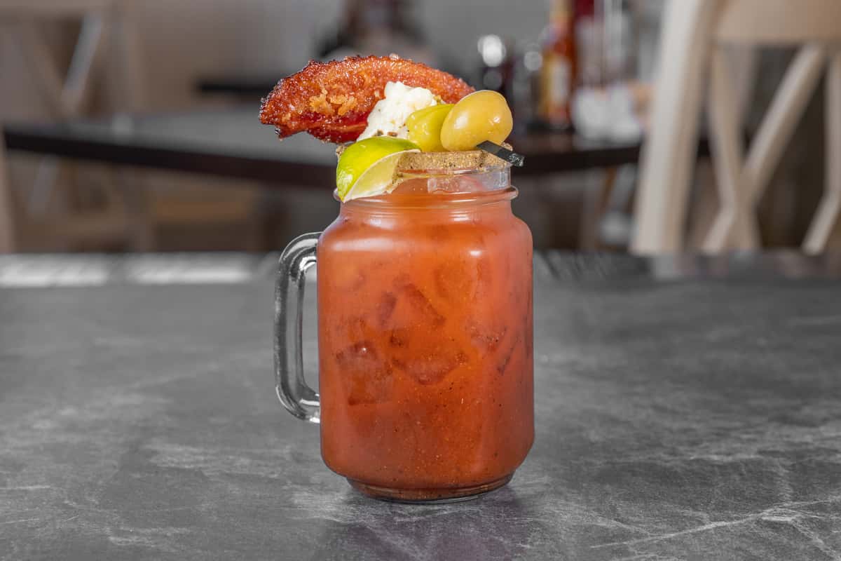 Spicy red cocktail served in a mason jar with a salted rim, garnished with a crispy chicken wing, pickle, and lime wedge, on a dark tabletop.