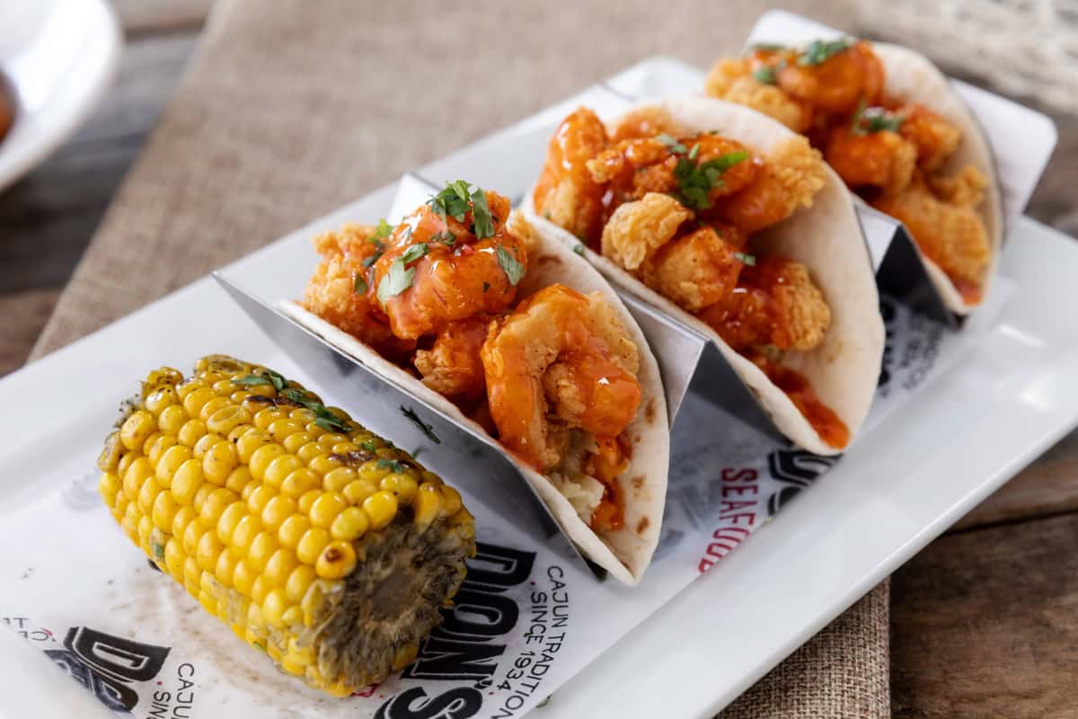 Crispy shrimp tacos drizzled with spicy orange sauce and topped with fresh herbs, served in soft tortillas alongside grilled corn on the cob on a casual seafood restaurant plate