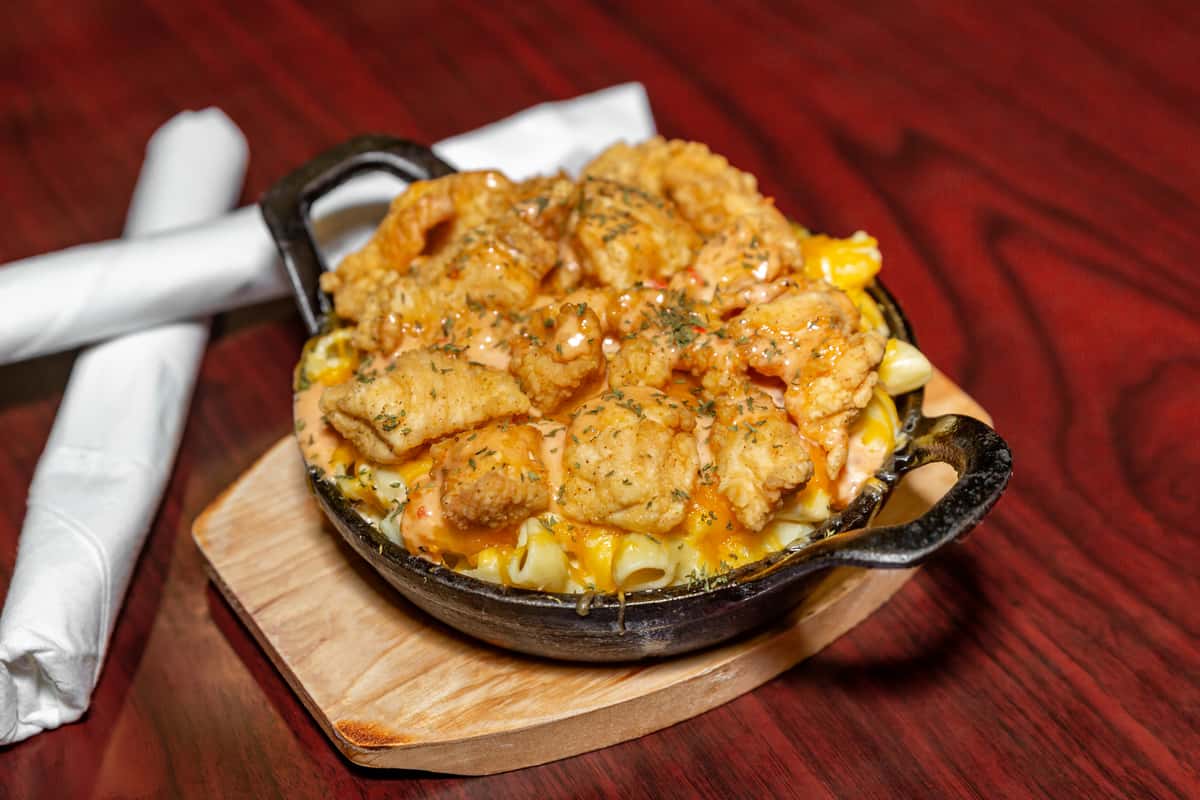 Skillet of creamy macaroni and cheese topped with crispy fried seafood and drizzled with sauce on a wooden serving board.
