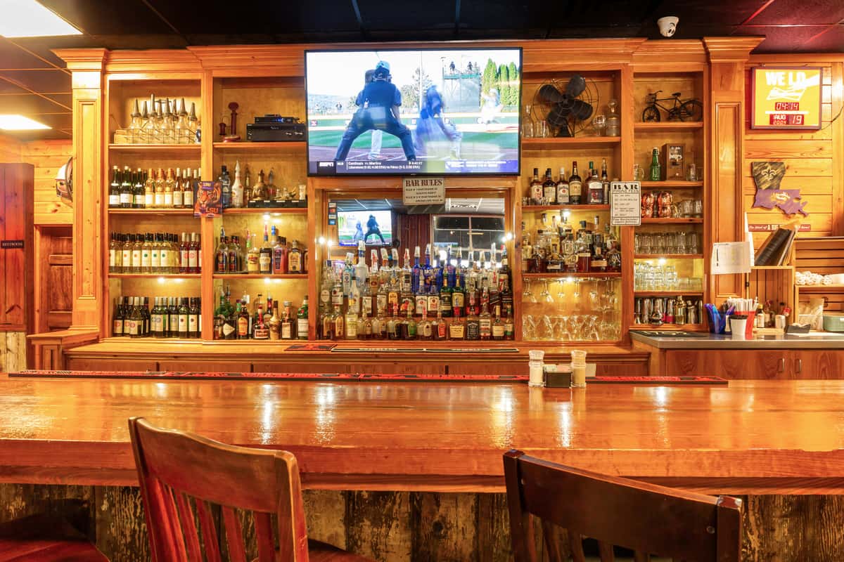 Well-stocked wooden bar with liquor bottles, glassware, and a TV showing a baseball game, viewed from across the polished counter.