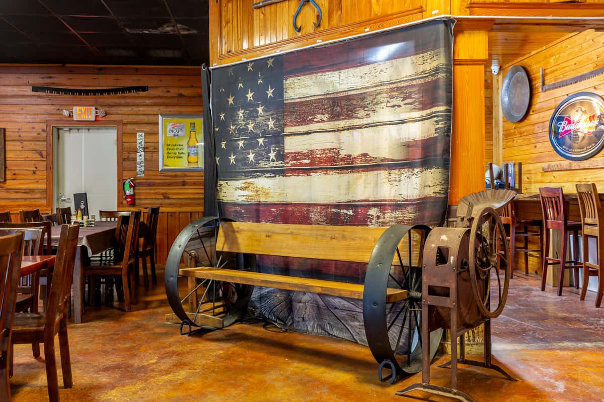 Wagon wheel bench with wooden seat in front of a vintage American flag backdrop inside a rustic wood-paneled restaurant.
