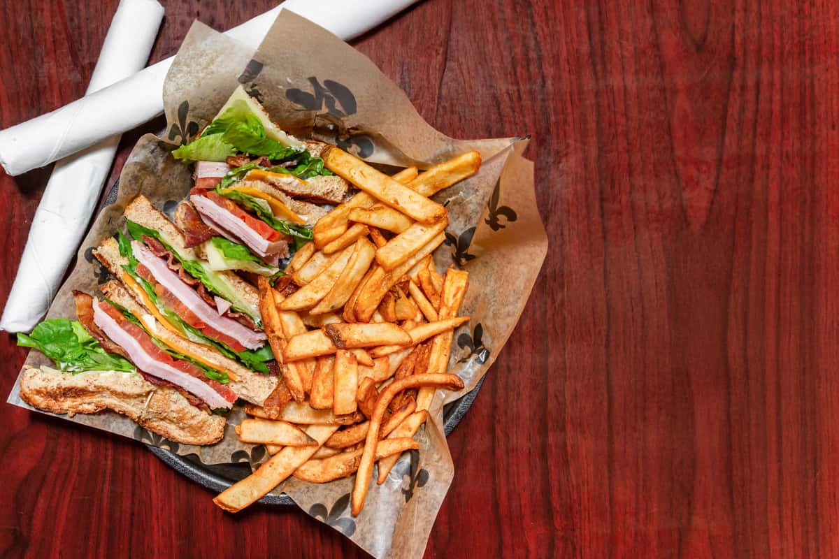 Club sandwich layered with ham, bacon, lettuce, tomato, and cheese, served with a side of golden French fries.