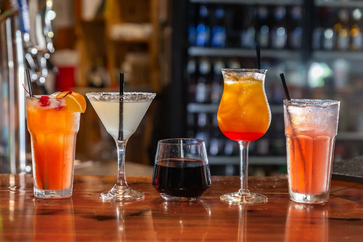 Variety of colorful cocktails and beverages lined up on a wooden bar counter with a blurred background of a bar setting.