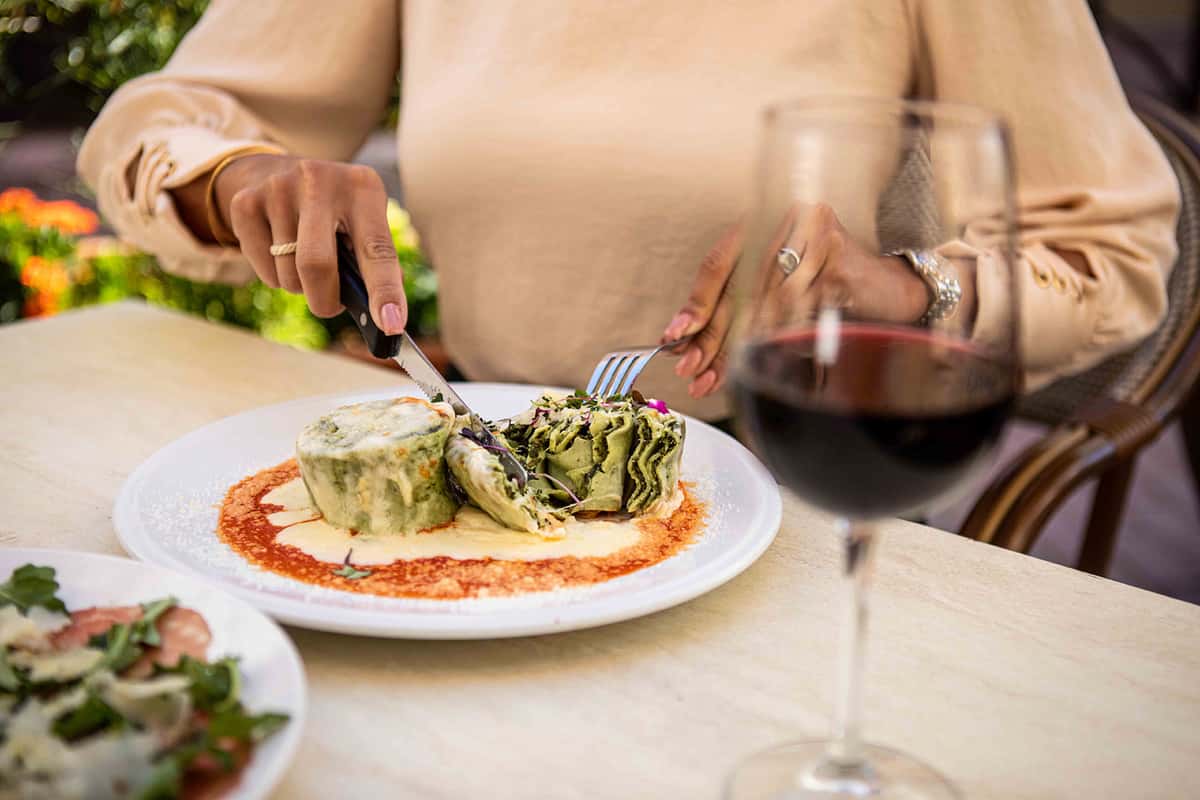 A beautifully plated Italian-style dish is being served at a dining table. A person is using utensils to cut into what appears to be stuffed pasta or a rolled entrée, topped with a creamy sauce, grated cheese, and herbs. The plate sits on a red sauce base, adding a vibrant contrast. In the foreground, a glass of red wine enhances the elegant dining atmosphere, while the softly blurred background suggests a cozy, upscale restaurant setting.