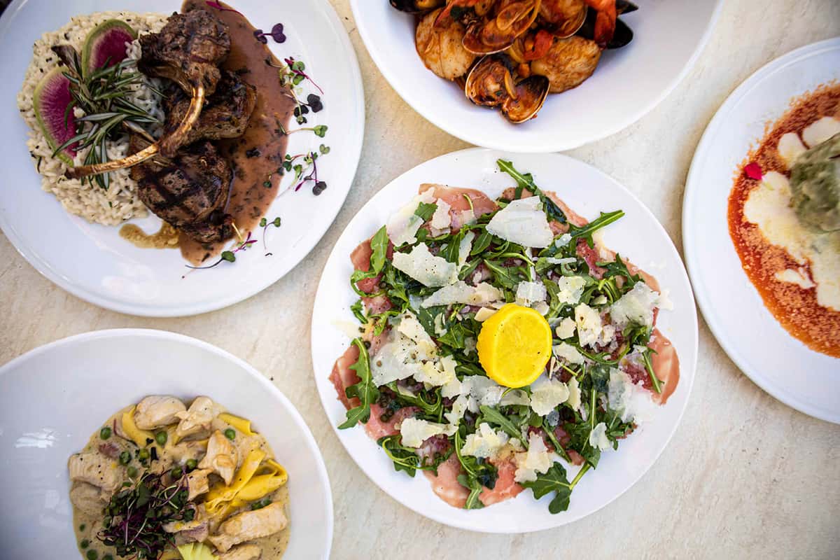 An overhead view of a vibrant spread of dishes, featuring a fresh salad topped with a bright egg yolk, alongside other plates of pasta and savory items. The presentation is colorful and abundant, showcasing a variety of textures and ingredients typical of a rich Italian dining experience.