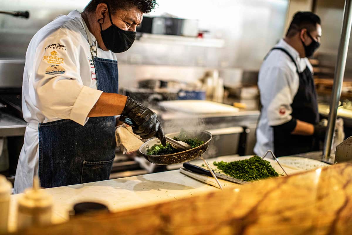 A chef, dressed in professional attire and wearing a face mask, skillfully plates a dish in the kitchen. Fresh ingredients are neatly arranged on the counter, highlighting attention to detail, freshness, and the artistry behind each meal.