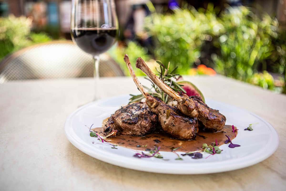 A beautifully plated rack of lamb is presented outdoors, perfectly cooked and arranged with the bones elegantly crossed. The dish is finished with a rich glaze and garnished with fresh herbs, while a glass of red wine sits nearby, enhancing the upscale, refined dining experience.