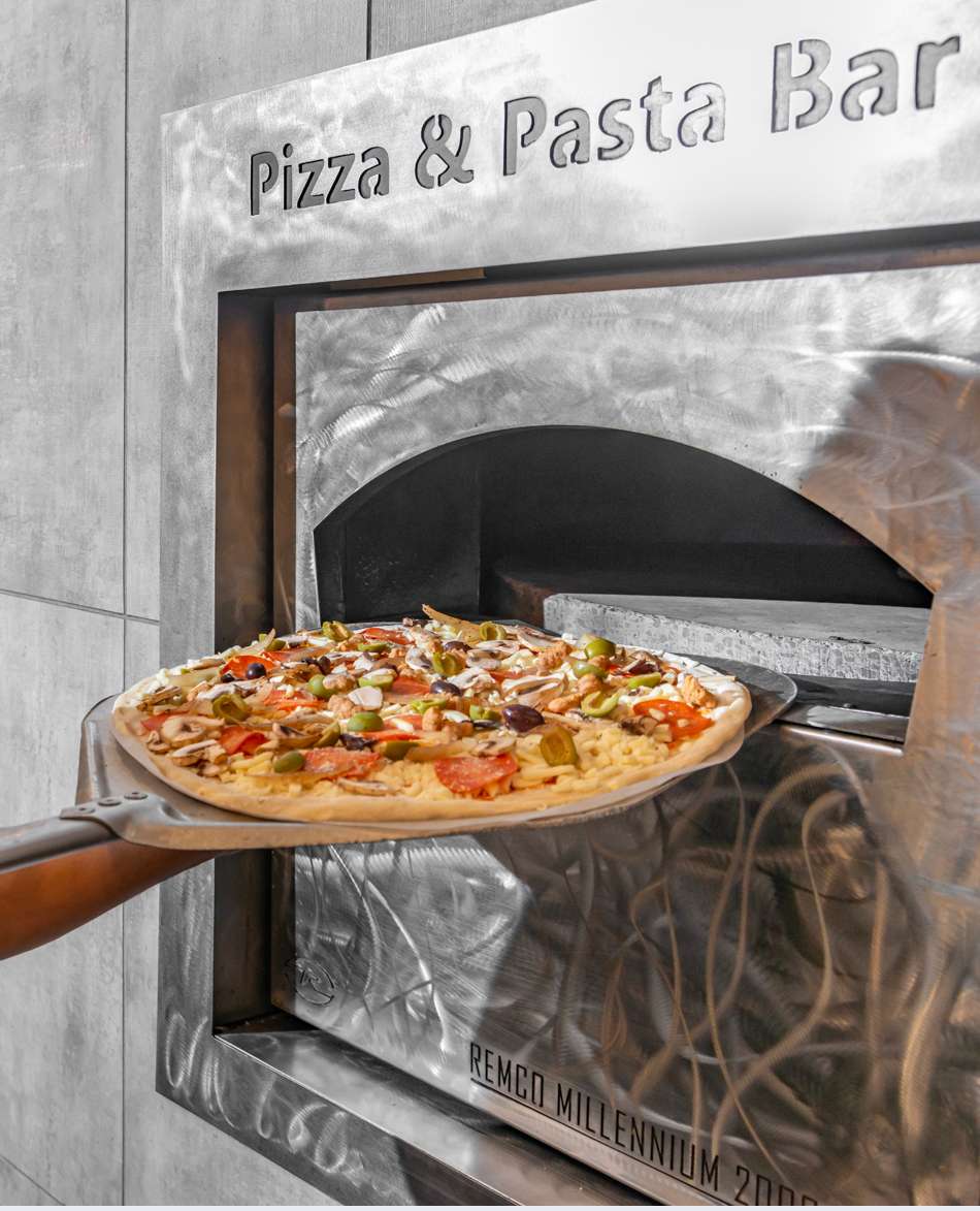 Wood-fired pizza with mushrooms, olives, and peppers being pulled from a stainless steel oven using a pizza peel.