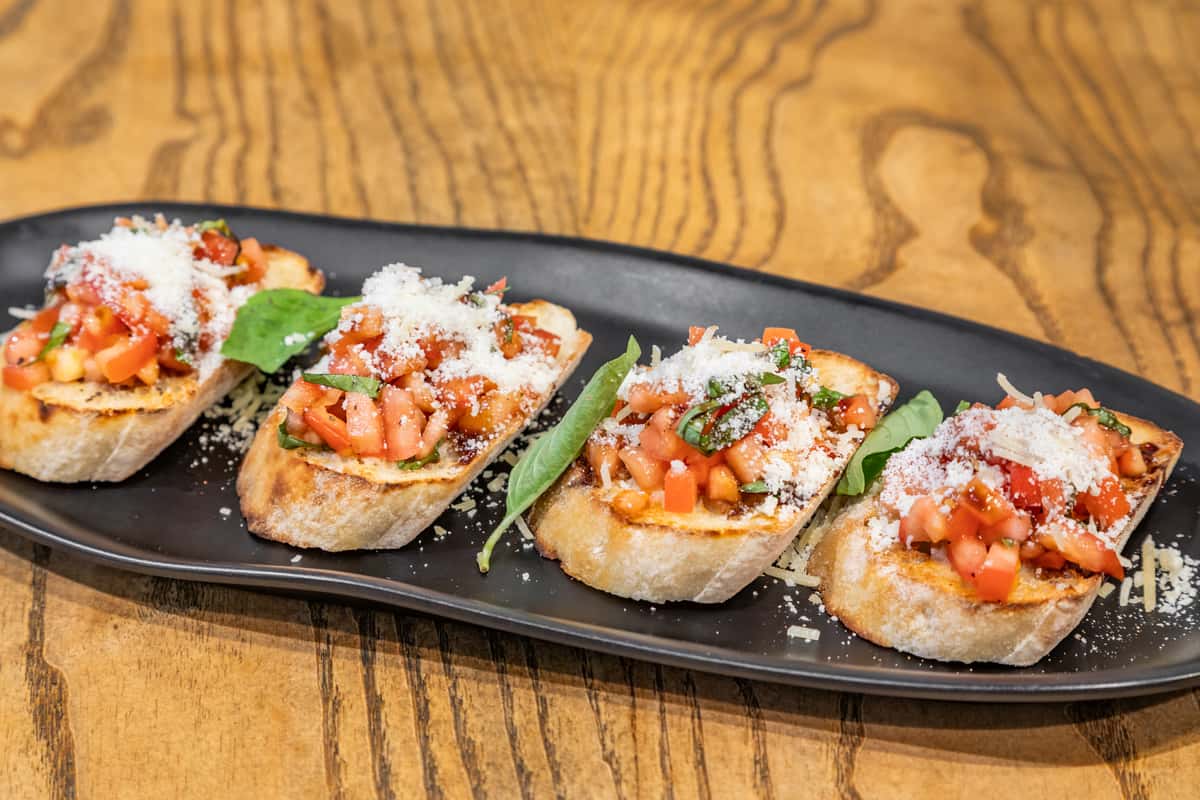 Toasted bruschetta topped with diced tomato, basil, and shredded parmesan, served on a black oval plate over a wood table.