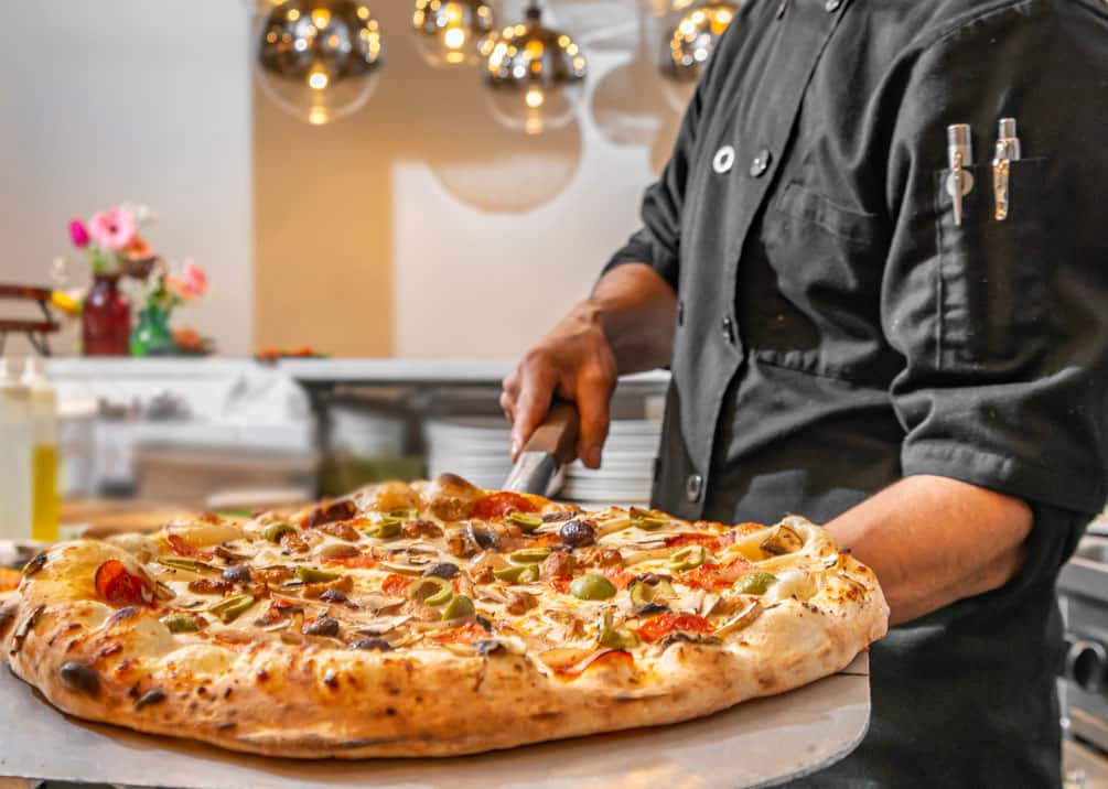 Chef holding freshly baked wood-fired pizza with olives, mushrooms, and artichokes in a modern kitchen.