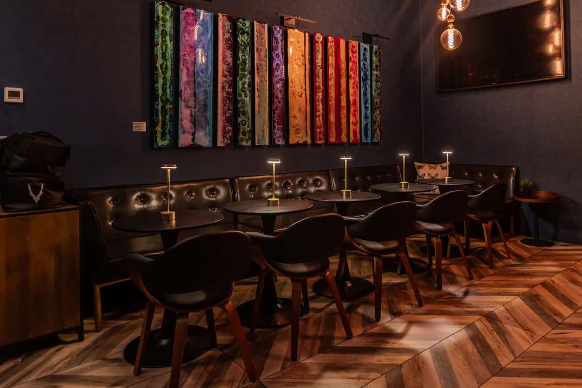 Chic restaurant lounge with a long tufted leather banquette, modern black chairs, and individual gold table lamps, accented by a colorful abstract wall installation and warm pendant lighting.