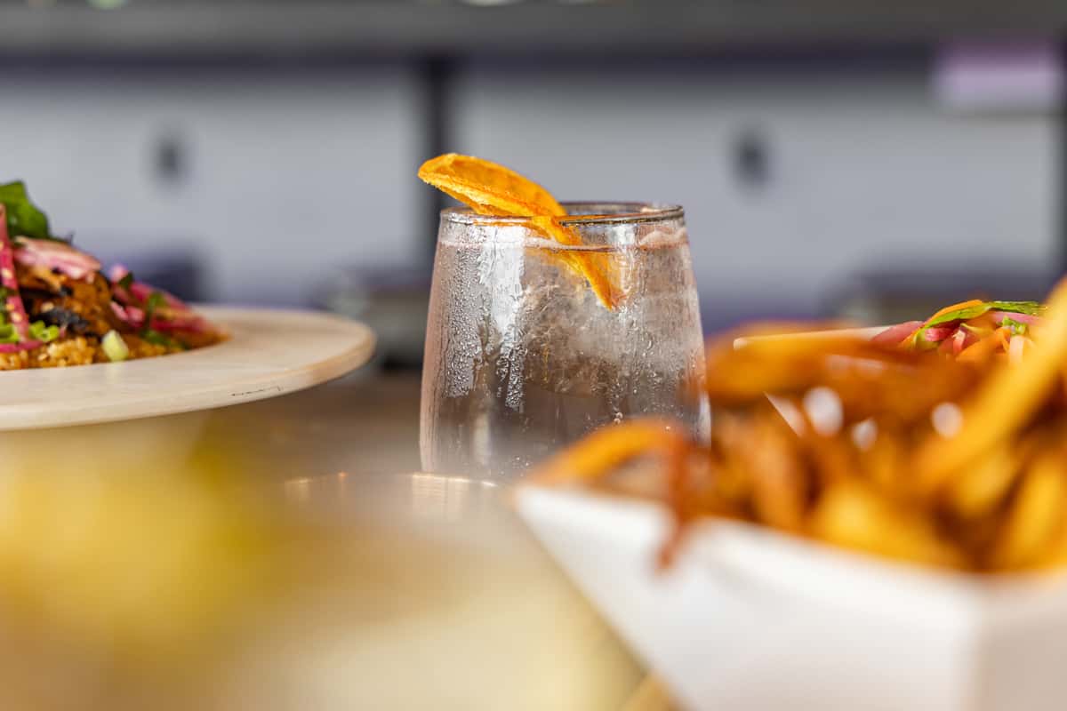 Chilled cocktail served in a clear glass with condensation, garnished with a dried orange slice, surrounded by plates of crispy fries and vibrant salad in a modern restaurant setting.