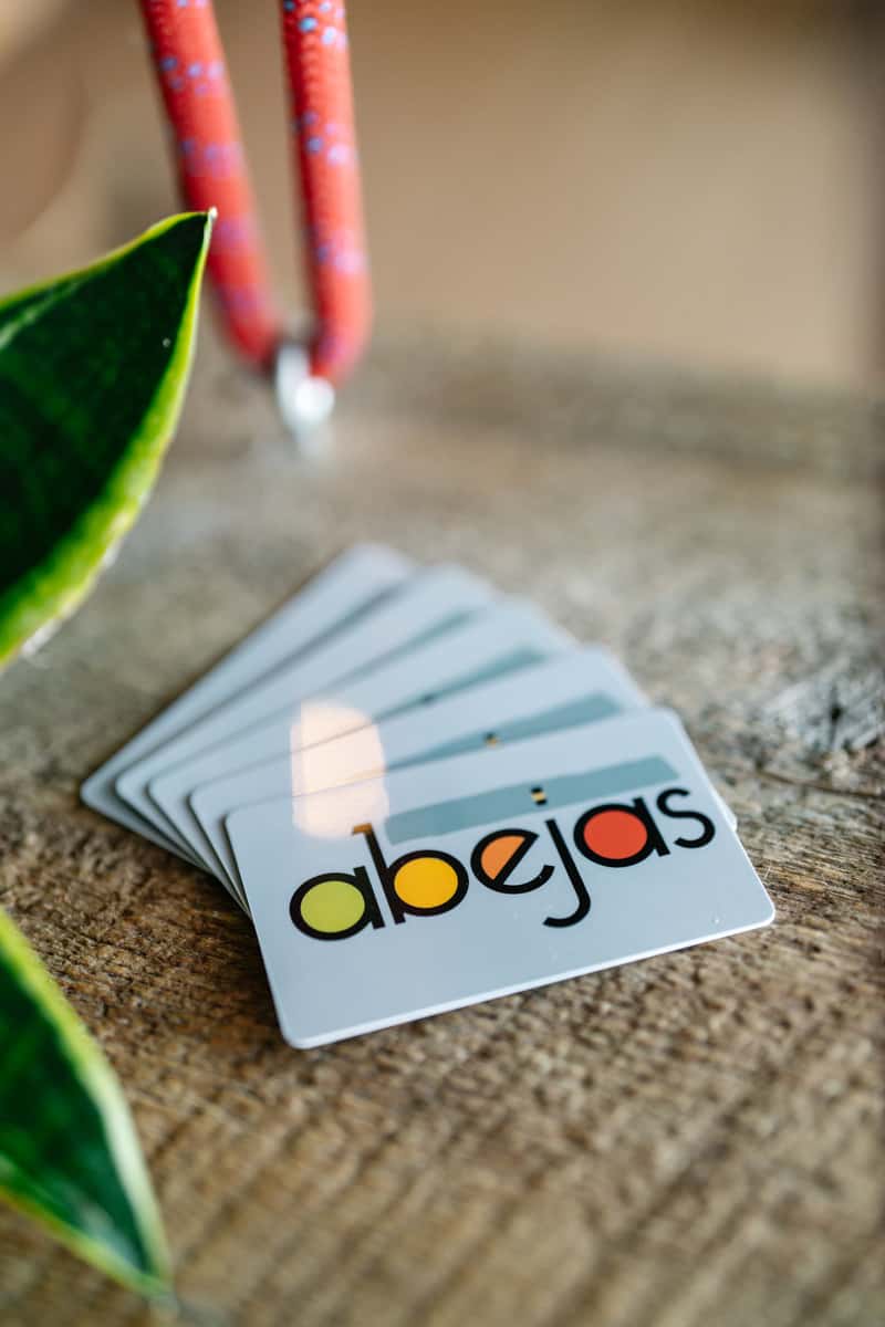 "Abejas" cards are stacked on a wooden surface next to plant leaves and a red cord. The word on the cards is spelled out with colorful letters.