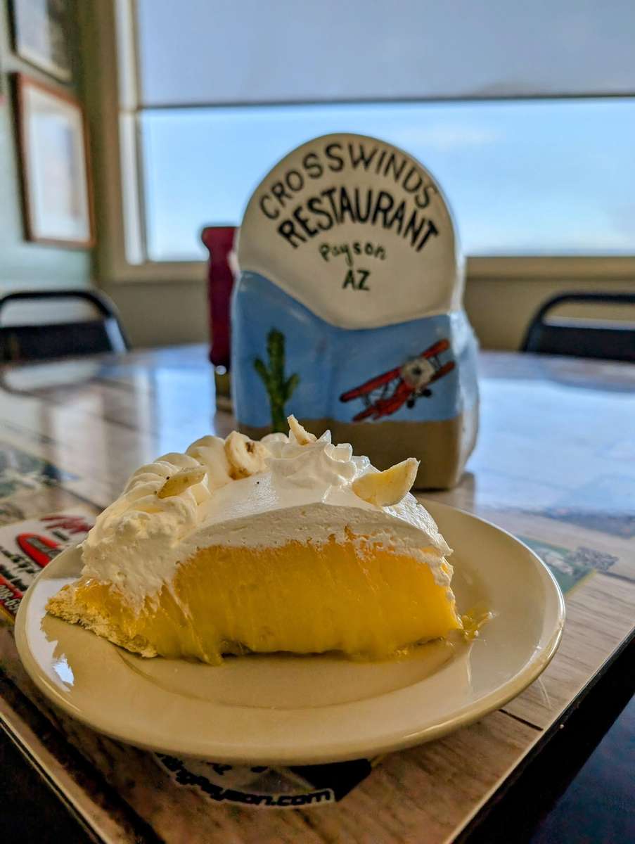 Slice of banana cream pie topped with whipped cream, served at Crosswinds Restaurant in Payson, AZ, with aviation-themed decor in the background.