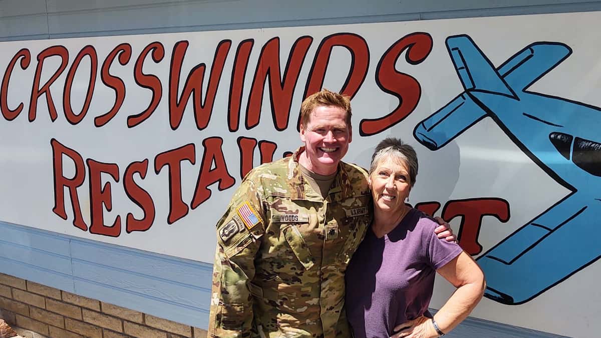A smiling U.S. Army service member poses with a woman in front of the Crosswinds Restaurant mural in Payson, AZ, featuring bold red text and a blue airplane illustration.