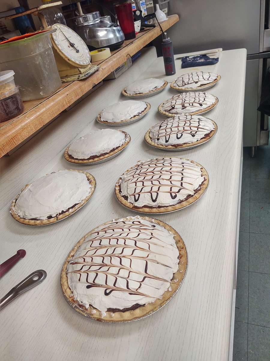 Row of freshly made chocolate cream pies topped with whipped cream, some drizzled with a chocolate lattice design, lined up on a prep counter at Crosswinds Restaurant in Sedona.