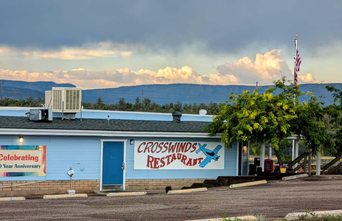 Exterior view of Crosswinds Restaurant in Payson, Arizona, featuring a bright blue building with a vintage airplane mural, set against a dramatic mountain backdrop under cloudy skies.