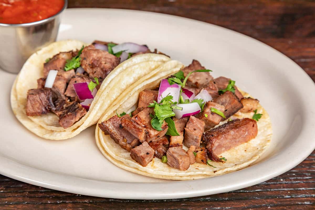 Two street-style carne asada tacos topped with diced red onions and fresh cilantro, served on double corn tortillas with a side of salsa.