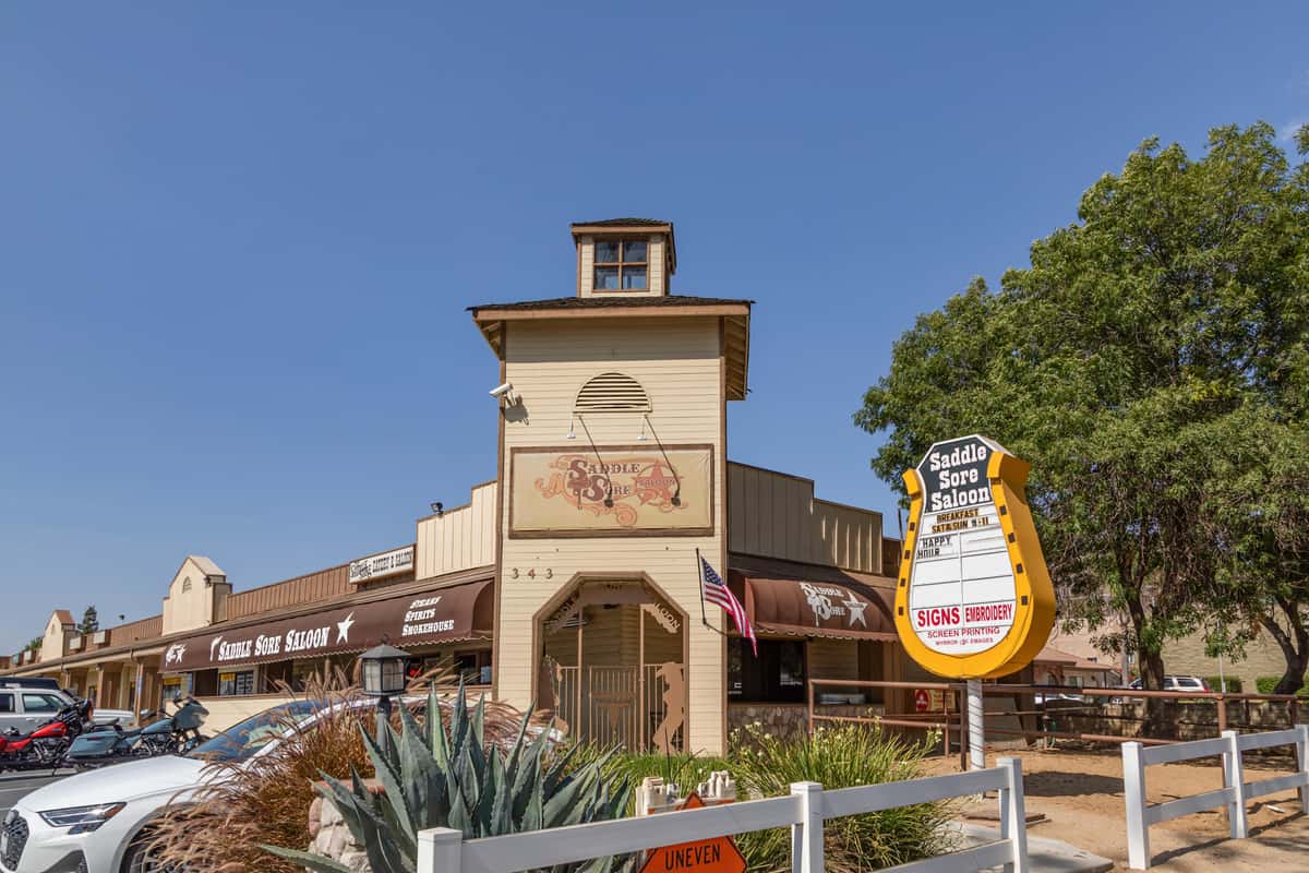 Front view of Saddle Sore Saloon with western-style architecture, outdoor sign, and parked vehicles on a sunny day.