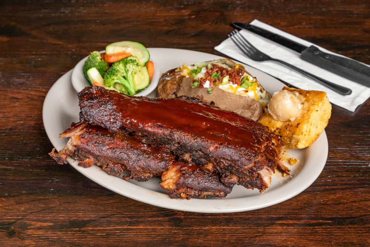Barbecue ribs glazed with sauce, served with steamed vegetables, loaded baked potato, and a slice of cornbread with butter.