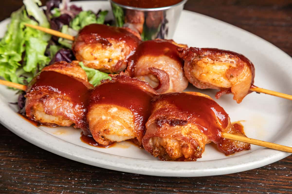 Bacon-wrapped shrimp skewers drizzled with barbecue sauce, served with fresh greens and a side of dipping sauce.