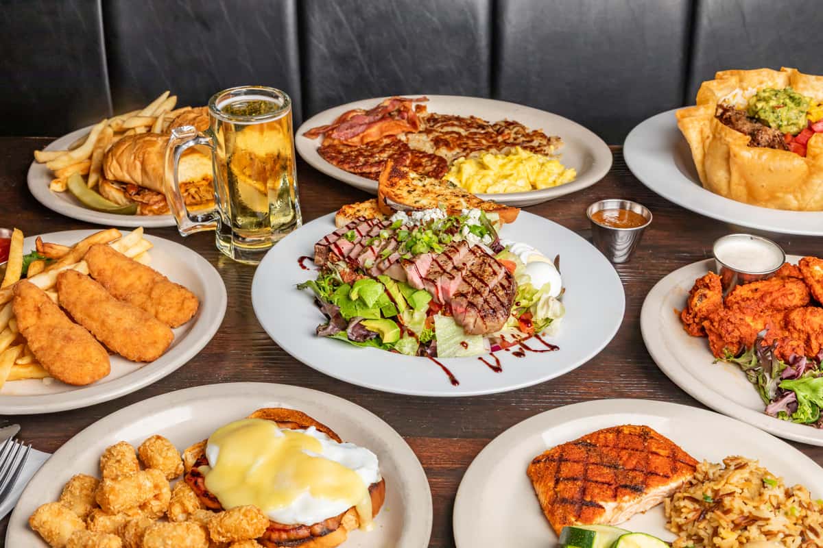 Assorted hearty meals including steak salad, fried chicken, tacos, breakfast plates, fish and chips, and a cold mug of beer.