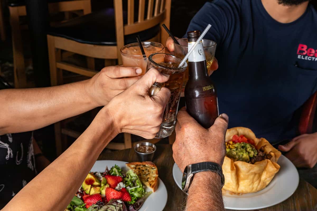 Friends toasting drinks over a table with fresh salads and a taco bowl, celebrating in a casual restaurant setting.