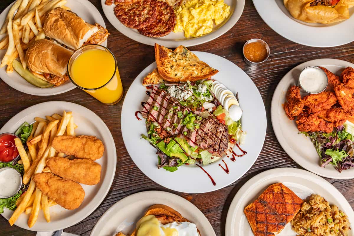 Assorted comfort foods including steak salad, fried chicken, fish and chips, breakfast platter, and orange juice on a wooden table.