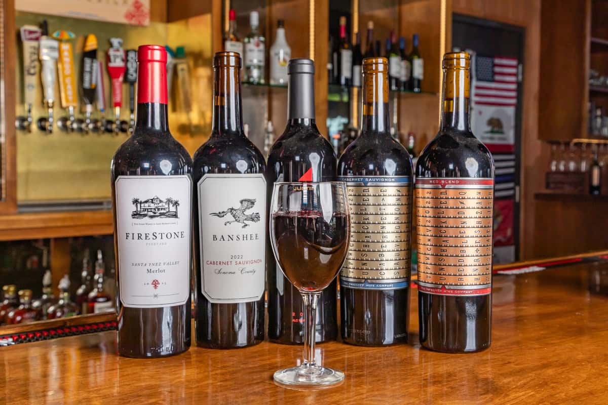Five bottles of red wine and a filled glass on a polished wooden bar, with taps and liquor bottles in the background.